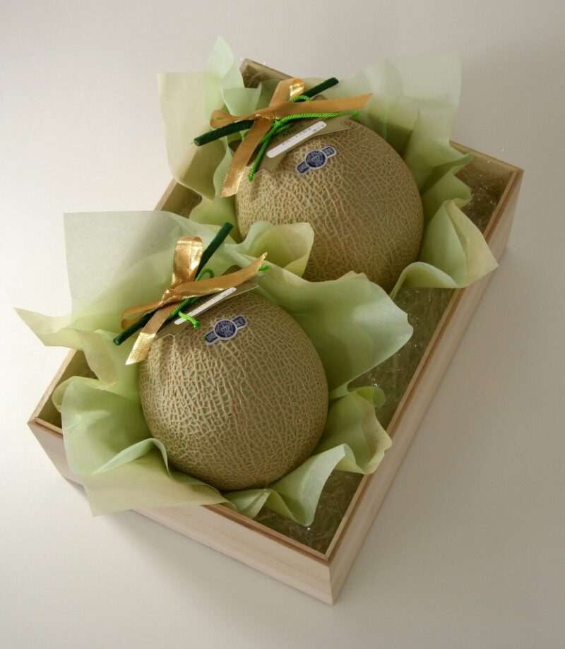 Two large melons in a wooden box placed on green paper as they are presented as Japanese omiyage