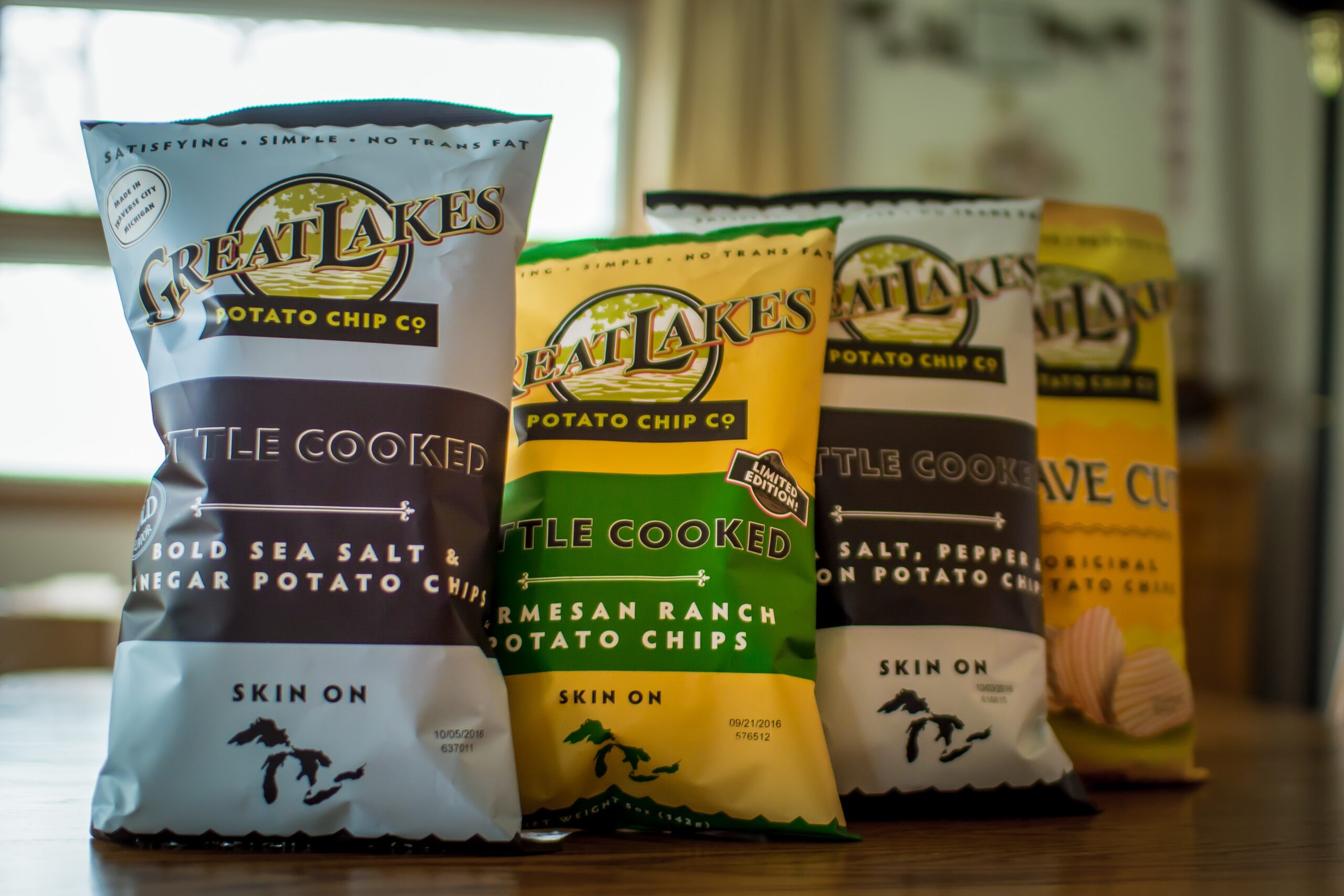 Four bags of potato chips proudly lines up on a table in house, alternating between a blue bag and a yellow bag