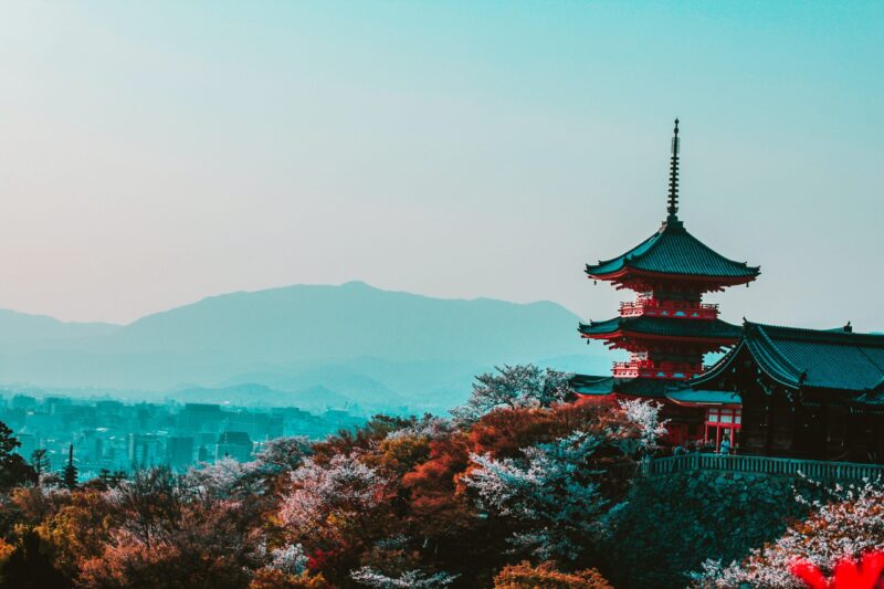 Japanese Temple peaking out of the tree line with a blue tinted mountain in the background located in Kyoto, a perfect place to find Japanese omiyage for loved ones