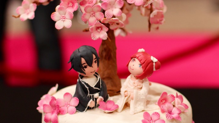 A cake decorated with a white fondant base, a Sakura tree with falling flowers, and a newly married couple kneeling underneath in their marriage attire