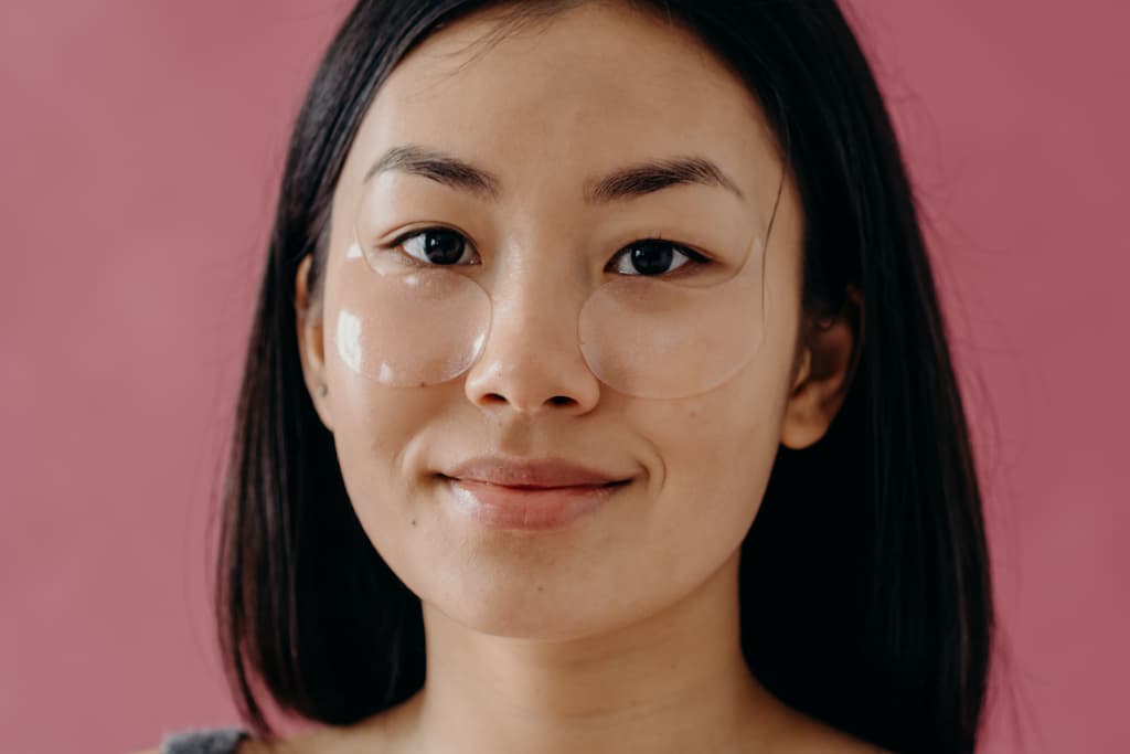 A young asian women is in front of a dusty pink bacjground as she gazes striaght at you with a relaxed smile and eye masks laying just underneath her eyes