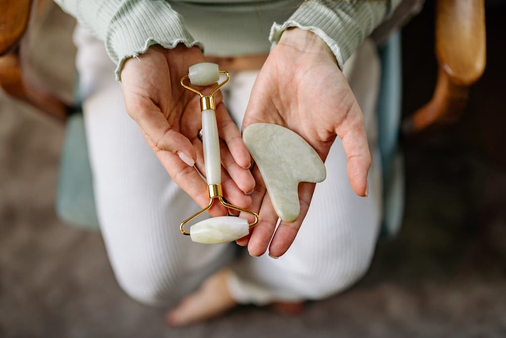 A woman kneels and has her hands open to show a jade roller and a jade color Guasha, whose use is a recommended step in the japanese skincare process