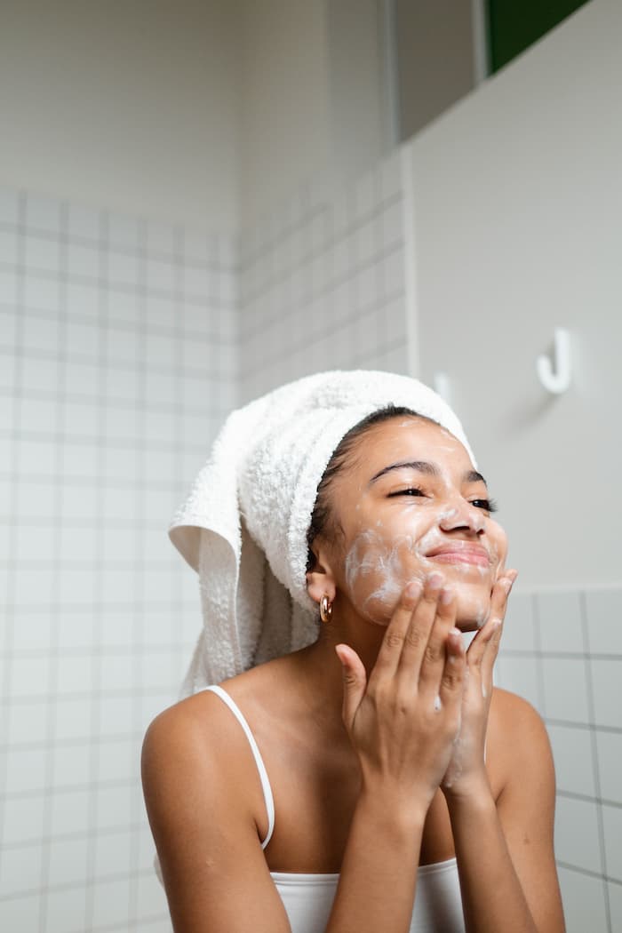 A young black women is in her bathroom wearing a white tank top with her hair wrapped up in a white towel as she washes her face and is smiling at herself in the mirror