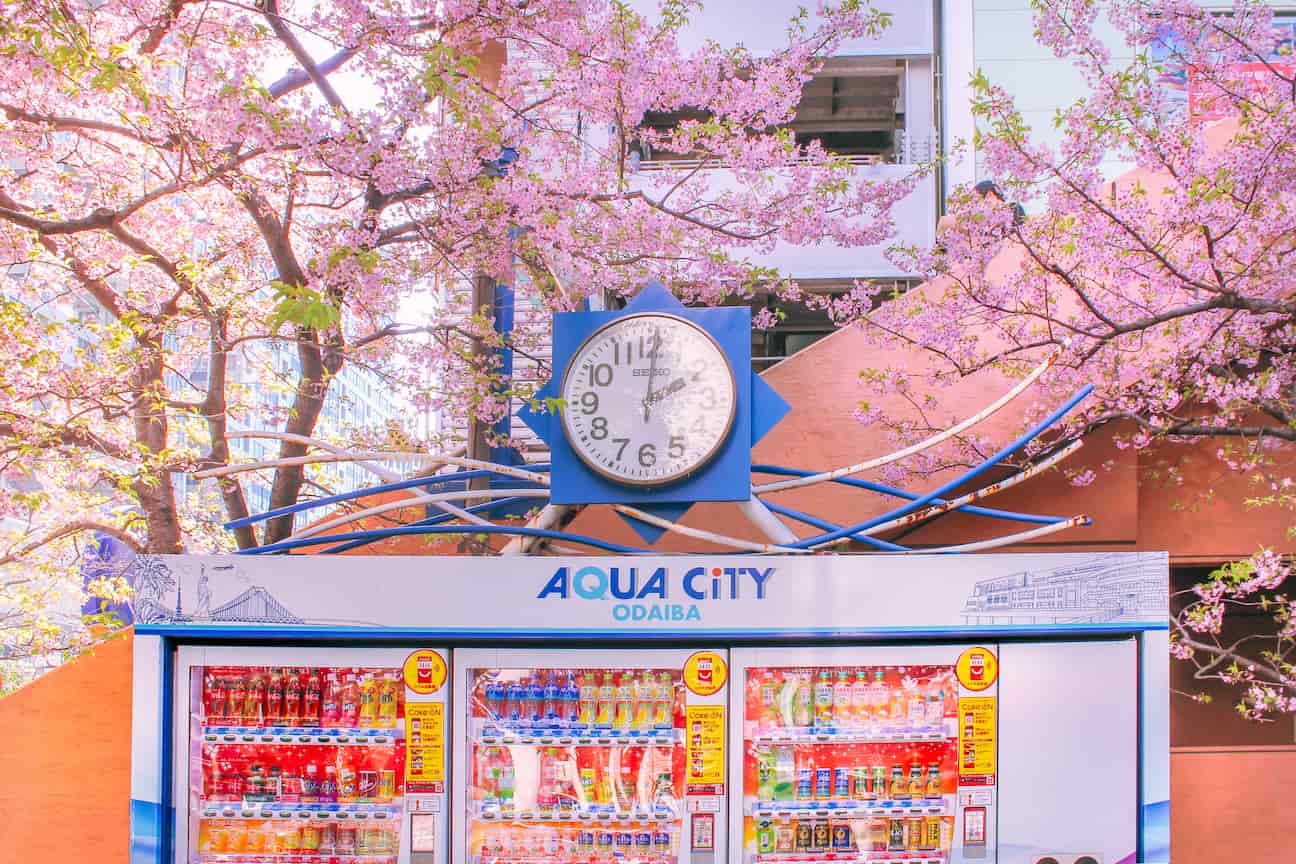 Several white vending machines lined up with a clock above and labelled Odaiba Aqua City