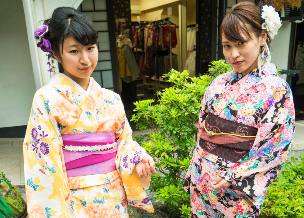 Two Japanese girls standing on the street wearing a yellow kimono with a pink obi (belt) and a pink kimono with a brown obi