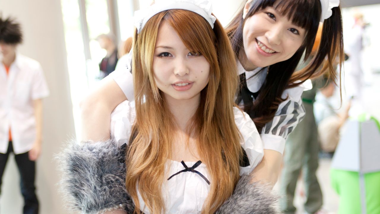 Two maids from a maid cafe in Tokyo - one has long brown hair and one has black hair. The one with black hair is hugging the other from behind