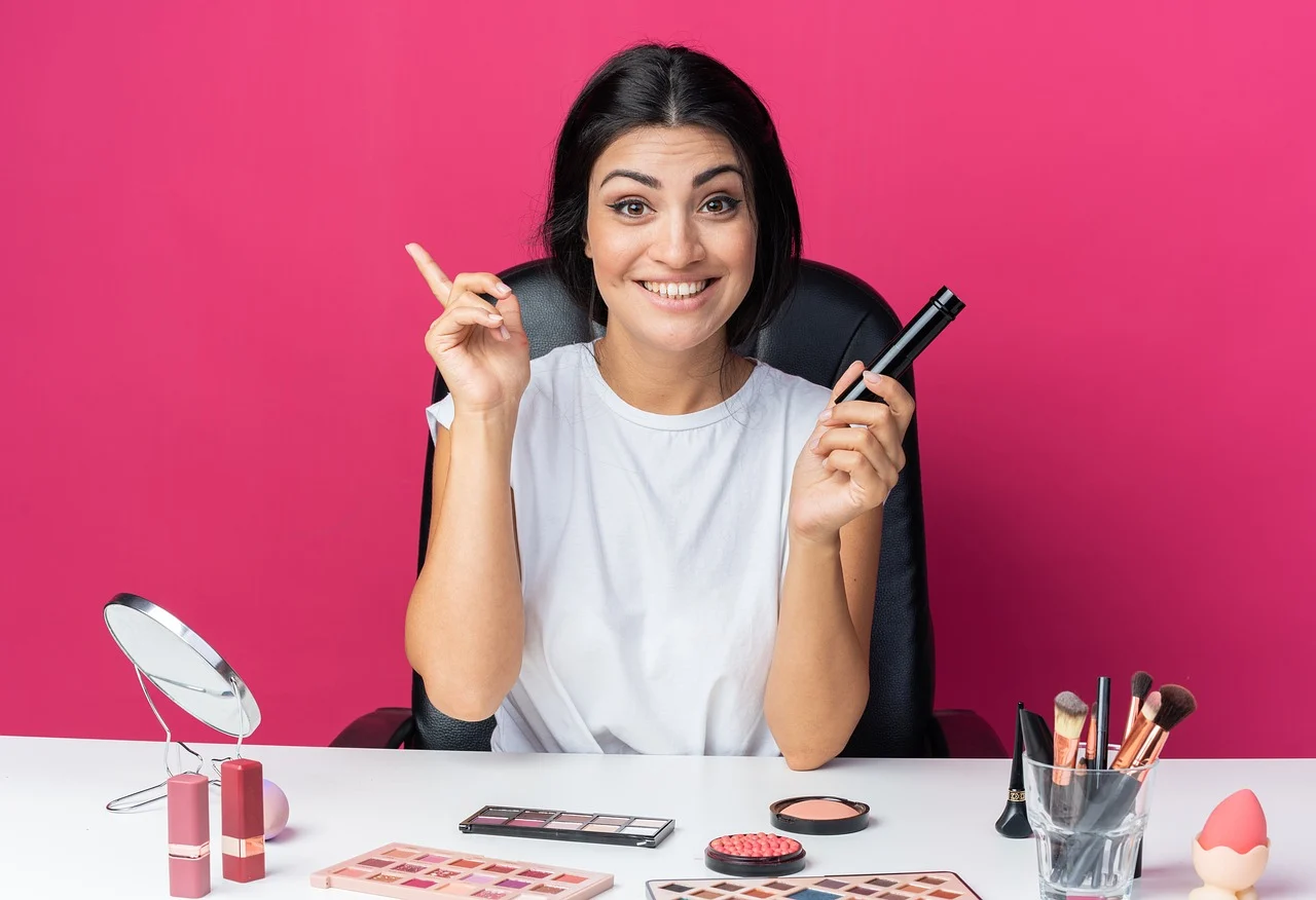 A lady wearing a white shirt with different Japanese makeup brands on the table in front of her while holding a lipstick and smiling