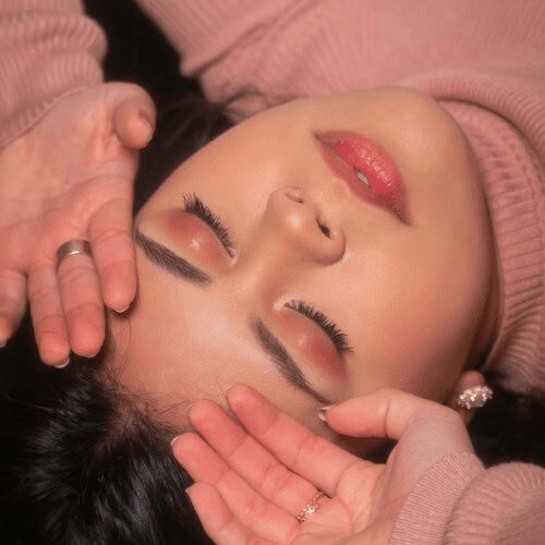 A face of a woman lying down with her hand on on the side of her forehead with her eyes closed revealing a bold eyeshadow on her eyes