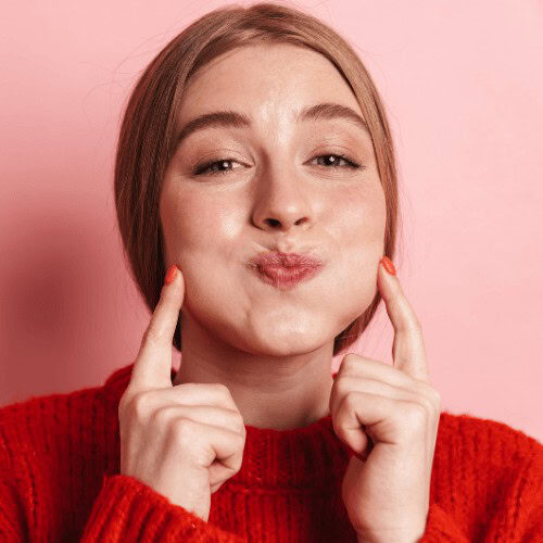 An image of a woman with pink background and red sweater with her lips pouting and her fingers pointing on both sides of her cheeks