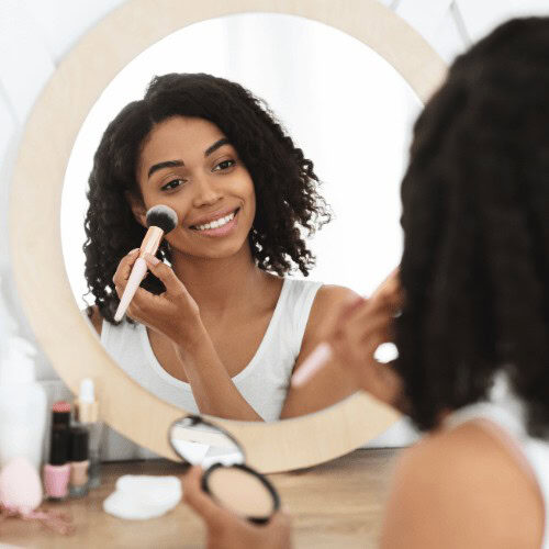 A woman looking at a beige, round mirror while putting a blush in her cheek using a brush and holding the cheek powder on her left hand