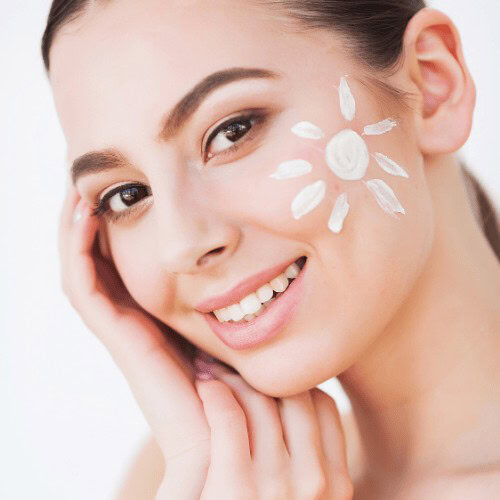 A face of a woman resting on her hand and smiling while looking to the camera with a spf cream on her cheeks that is shaped like a sun